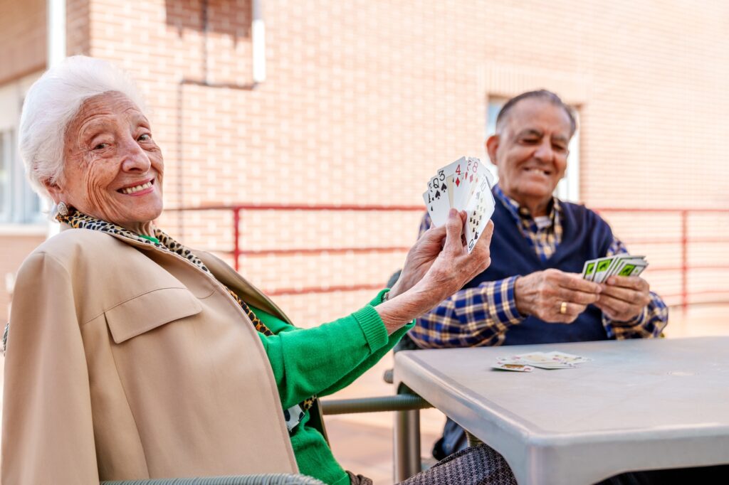 In the sunny geriatric patio, a cheerful elderly couple engage in a card game, exuding joy and contentment amidst the vibrant ambiance.