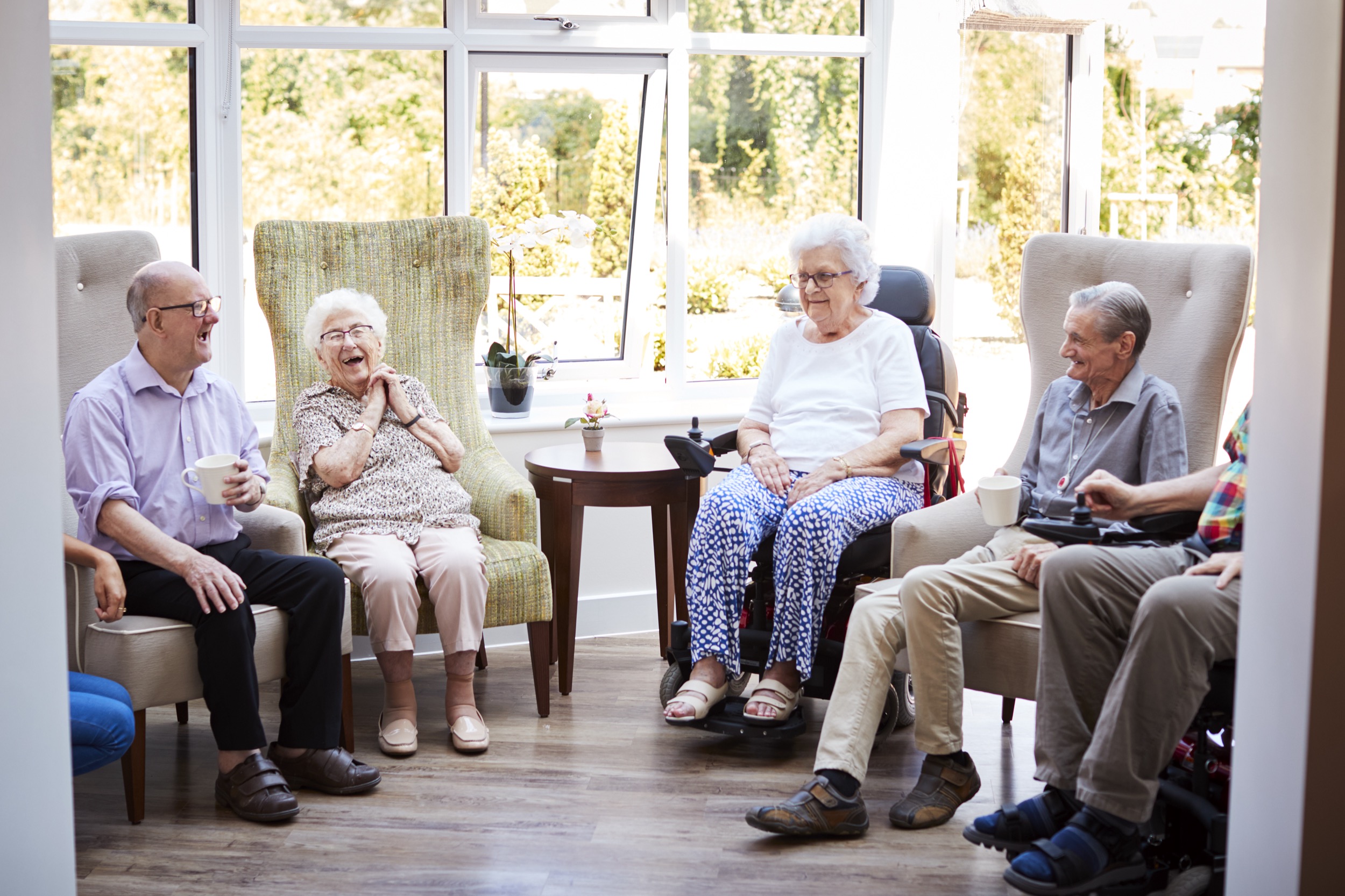 Male And Female Residents Sitting In Chairs And Talking In Lounge Of Retirement Home